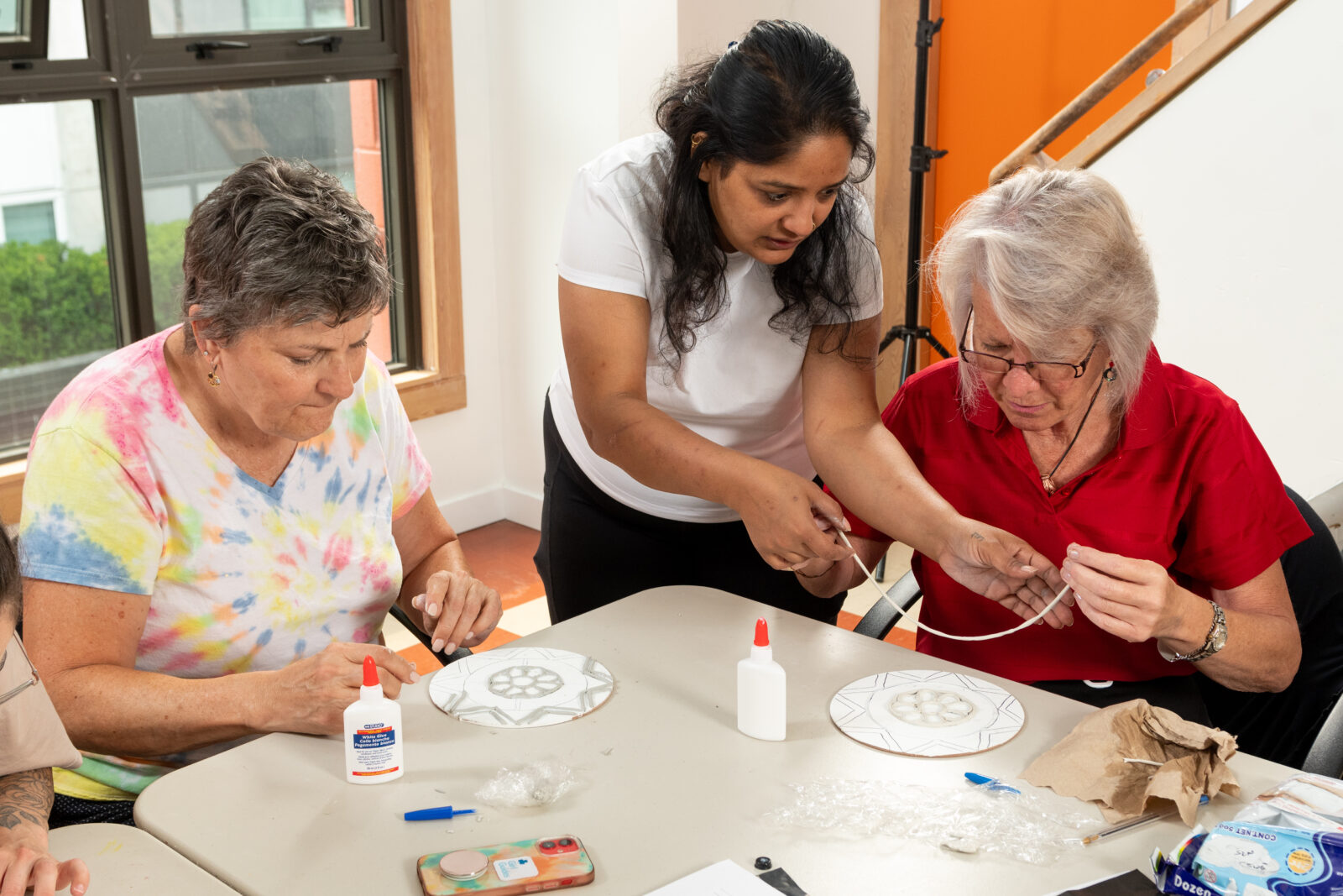 A volunteer assisting two senior women in an arts and crafts activity at Gordon Neighbourhood House. The women are focused on creating designs using glue and materials.