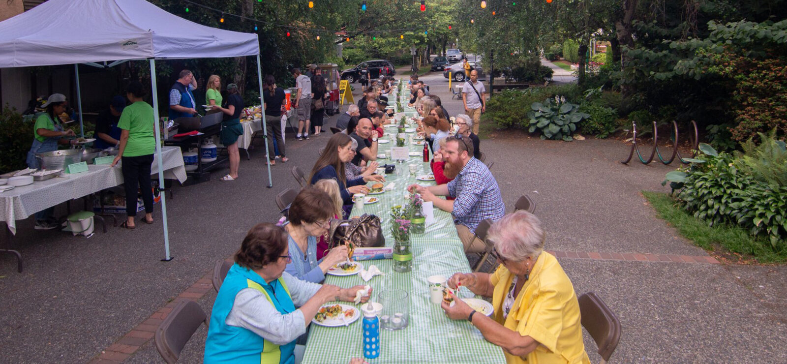An image of many community members gathered together to share a delicious meal at Gordon Neighbourhood House. The community members sit at a very long table in the plaza just outside of Gordon Neighbourhood House. You can see many different people of all ages enjoying a meal together. On the left, people in green t-shirts serve food to the community members. There are lights set up above the table for ambience.