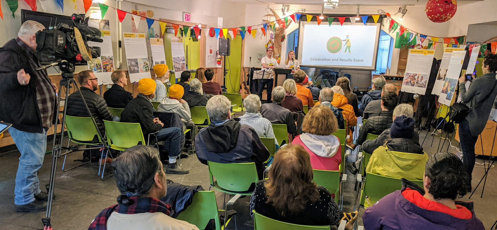 An image of a presentation at Gordon Neighbourhood House. Two people in white t-shirts stand in front of a projector screen that is showing a cartoon image of a man rolling a giant wheel, with the caption "Celebration and Results Event". Dozens of people sit in the audience facing the speakers. The audience is surrounded by multiple posterboards documenting the results that the speakers are talking about. A camera man at the back of the room films the audience and speakers.