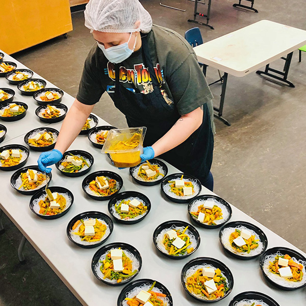 An image of a volunteer at Gordon Neighbourhood House putting together takeaway containers of a delicious meal. The person is wearing a white hairnet, a blue mask, a green t-shirt, and a black apron. They are spooning curry sauce over a bowl full of mixed vegetables and white rice. They are standing in front of a table full of takeaway bowls of curry, veggies, and white rice. These meals were delivered to seniors during the COVID-19 pandemic who experienced challenges accessing food.